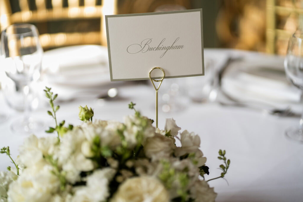 A white floral centrepiece with a card labelled Buckingham on a table set with glassware and plates, suggesting a formal event or wedding. David Bostock Photography