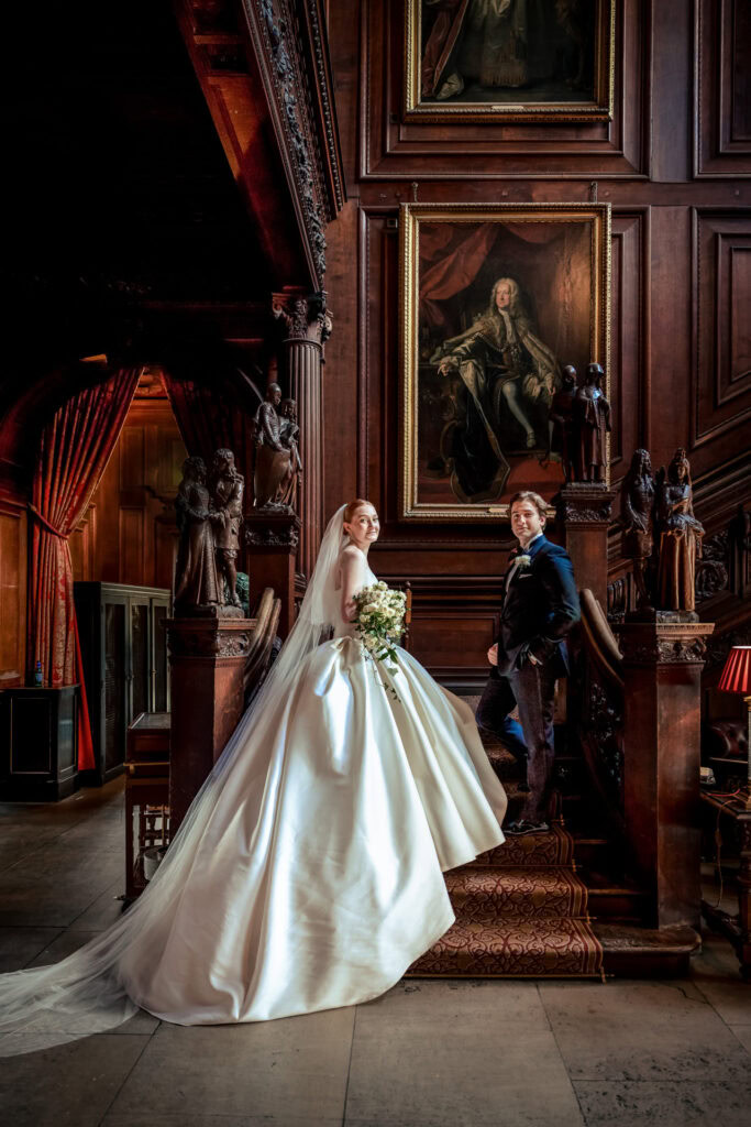 A bride in a white dress and a groom in a dark suit pose on an ornate wooden staircase with statues and a large framed portrait in the background of an elegant, historic interior. David Bostock Photography