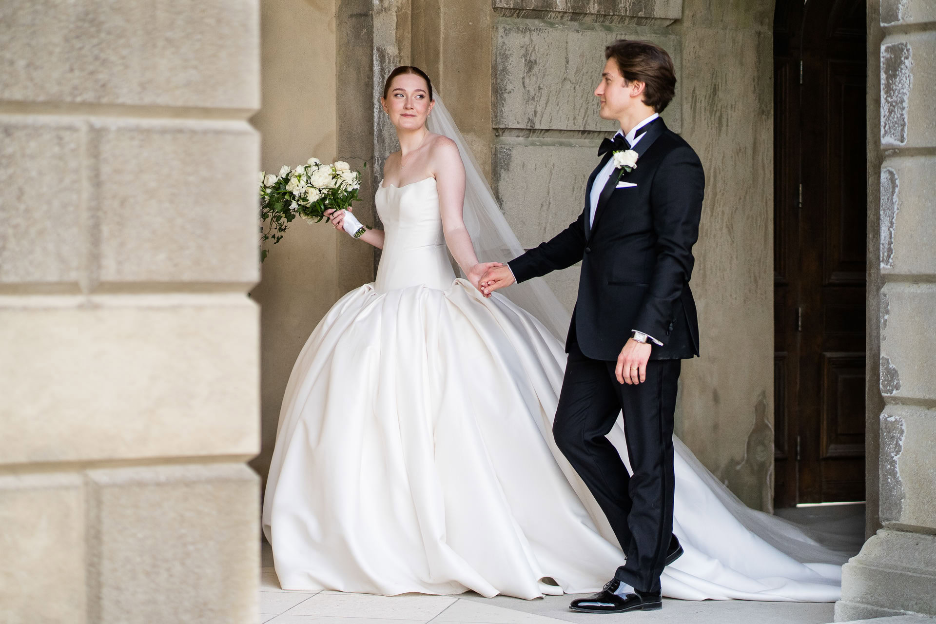 A bride in a white dress holds a bouquet and looks ahead, while a groom in a black dinner suit holds her hand and looks at her. They stand near stone columns and an arched doorway. David Bostock Photography