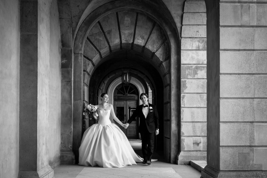 A bride in a full gown and a groom in a tuxedo hold hands, walking under a grand stone archway. The black and white photo captures an elegant, formal wedding moment. David Bostock Photography
