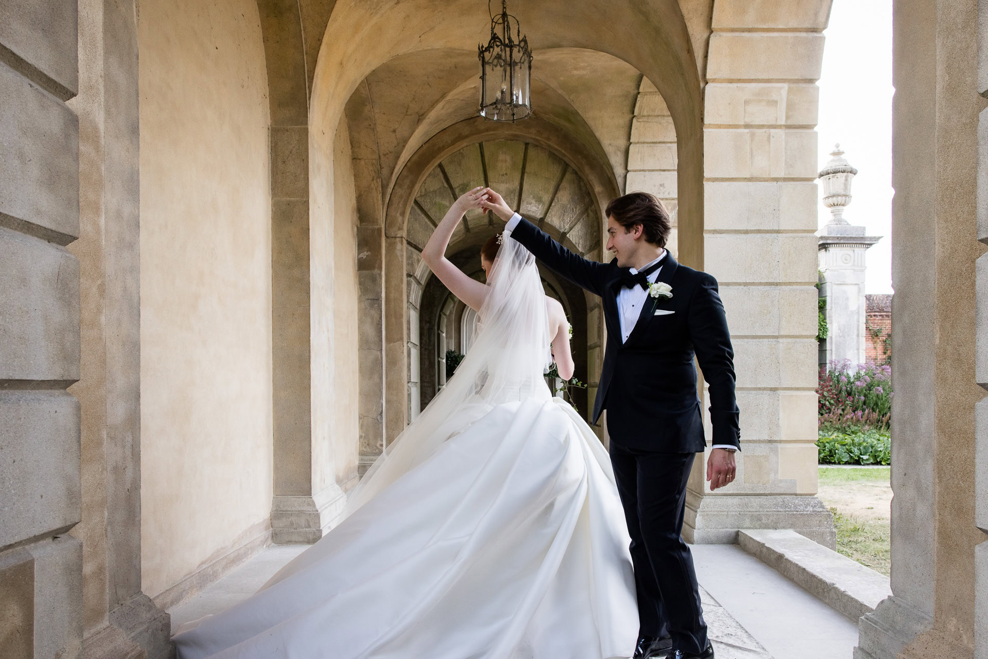 A bride and groom dance under stone arches. The groom, in a black tuxedo, twirls the bride, who wears a white dress and veil. Sunlight filters through the arches, creating a romantic atmosphere. David Bostock Photography