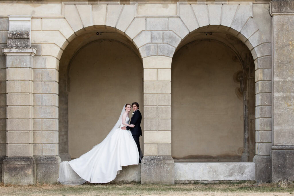 A bride in a white dress and a groom in a suit stand closely together under a stone archway of an old building, posing for a wedding photo. David Bostock Photography