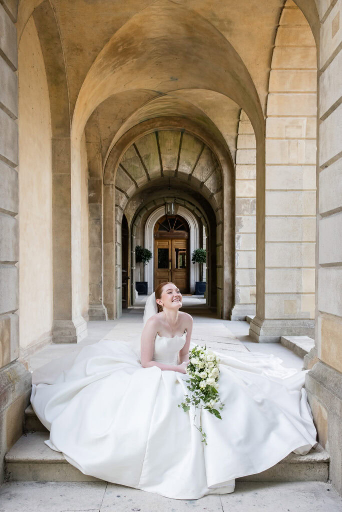 A bride in a white gown sits smiling under grand stone arches, holding a white and green bouquet, with her train spread around her on the floor. David Bostock Photography