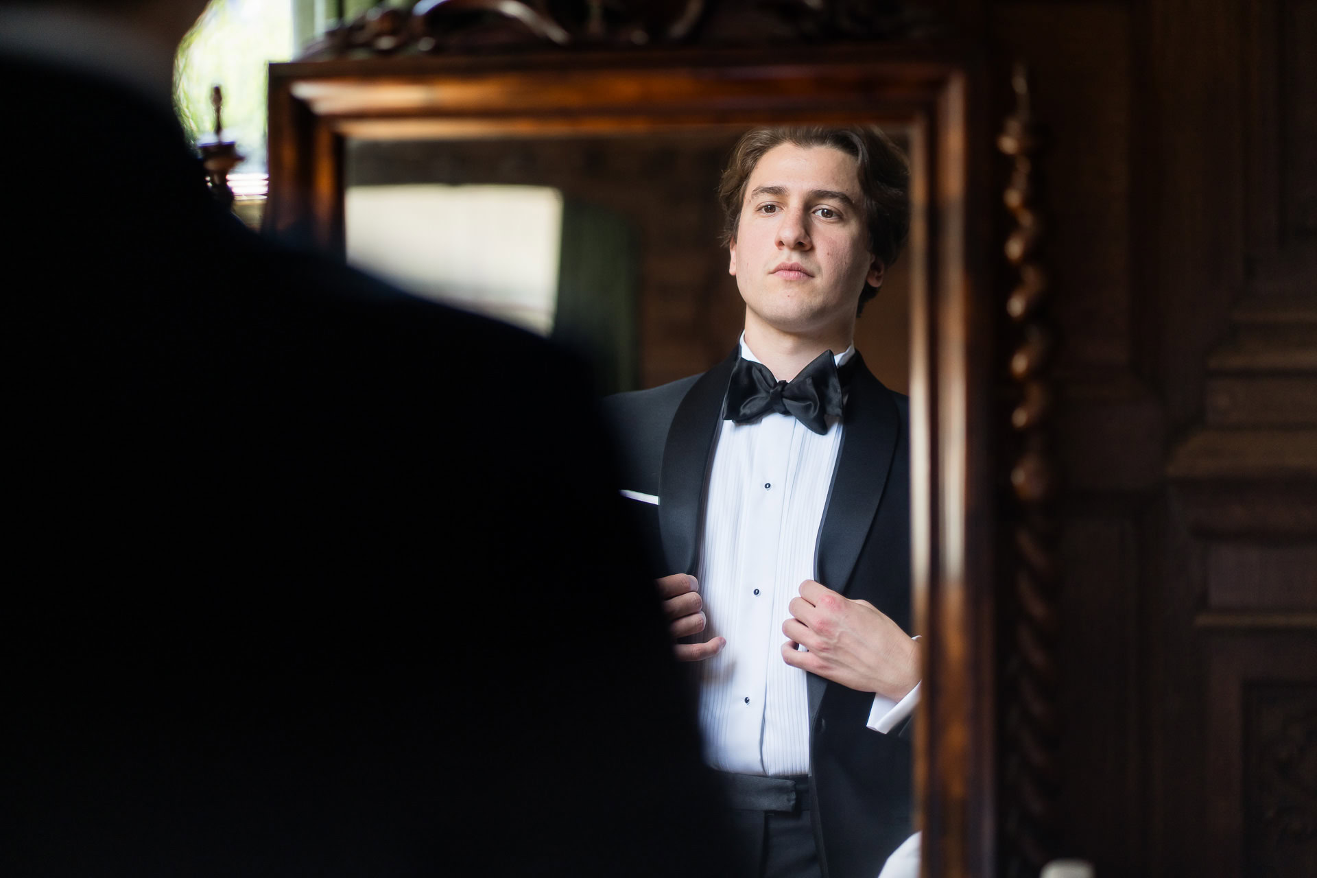 A man in a dinner jacket adjusts his jacket and looks confidently at himself in an ornate wooden mirror, with his reflection clearly visible. David Bostock Photography