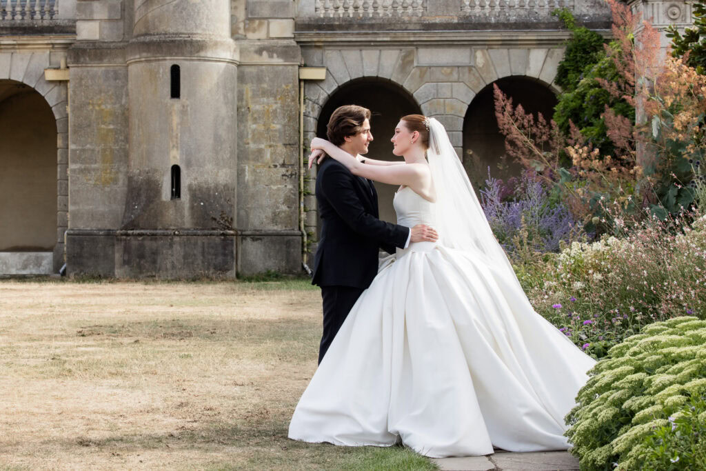 A bride and groom embrace outside a grand stone building, surrounded by colourful flowers and greenery. The bride wears a white dress and veil, while the groom wears a black suit. David Bostock Photography