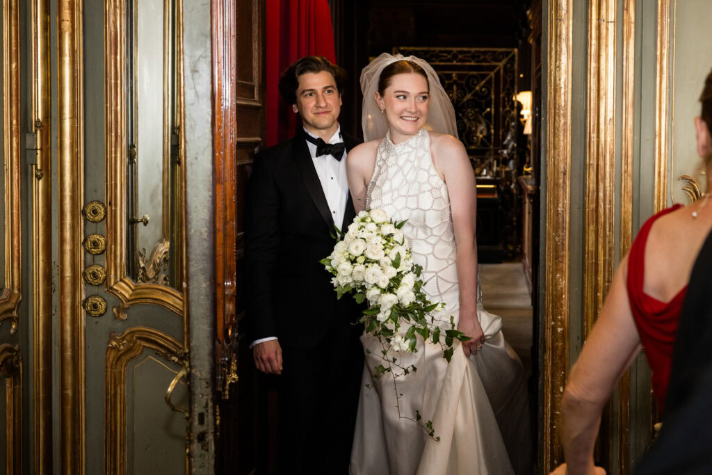A bride in a white dress holding a bouquet stands beside a groom in a tuxedo. They are smiling, framed by ornate golden doors, and facing a person in a red dress. David Bostock Photography