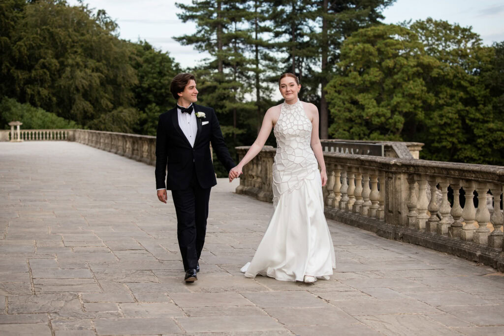 A bride in a white dress and a groom in a black dinner suit hold hands and walk outdoors on a stone pathway, surrounded by trees and greenery. David Bostock Photography