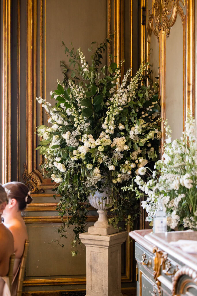 Large ornate vase filled with lush white flowers and greenery, set against a gold-trimmed wall in an elegant room; part of a formal gathering with seated guests visible. David Bostock Photography