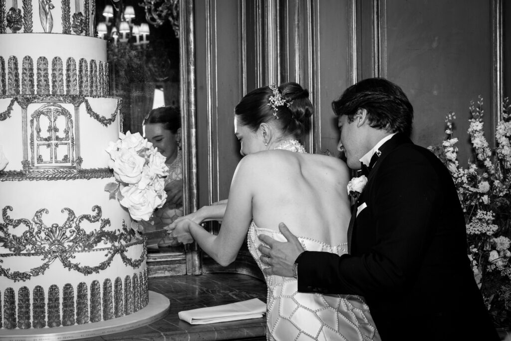 A bride and groom cut a large, ornate wedding cake together. The bride wears a strapless dress and the groom is in a suit, both focused on slicing the cake. Elegant décor and flowers are visible. David Bostock Photography