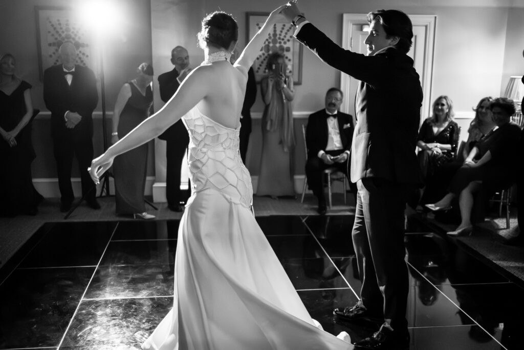 A couple in formal attire dances on a shiny floor at a wedding reception, surrounded by seated and standing guests watching them. The scene is in black and white. David Bostock Photography
