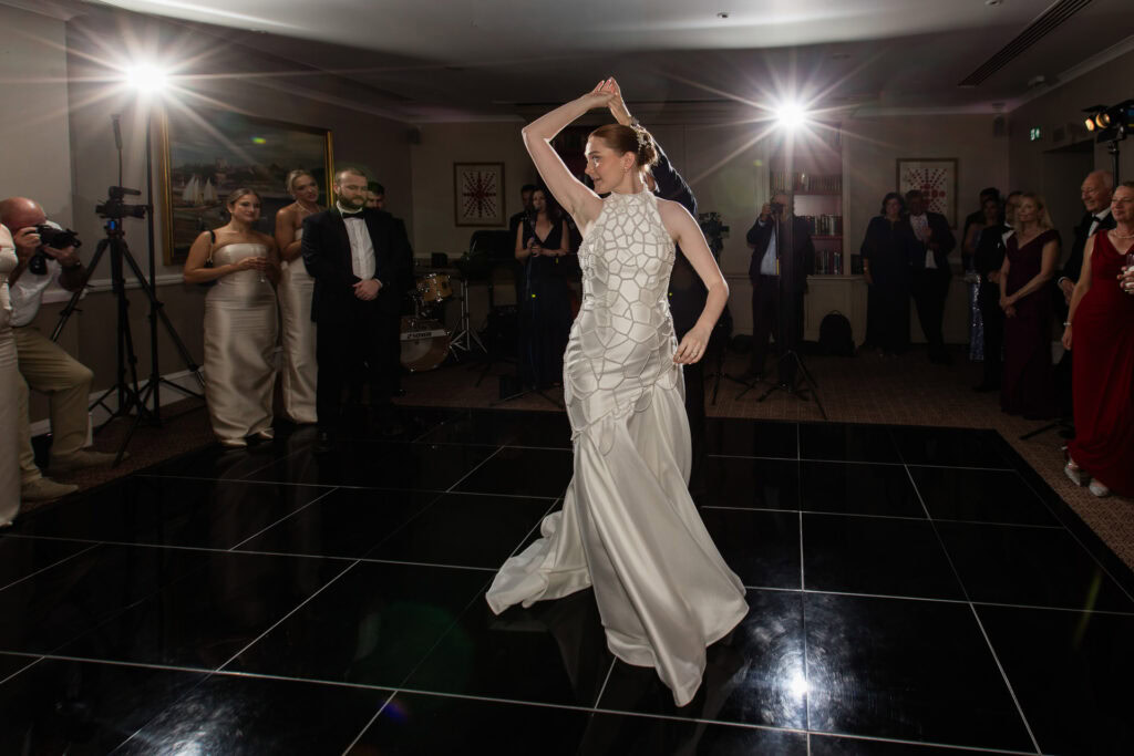 A woman in a white dress dances solo on a black-tiled floor, surrounded by elegantly dressed guests and photographers in a dimly lit room with bright spotlights. David Bostock Photography