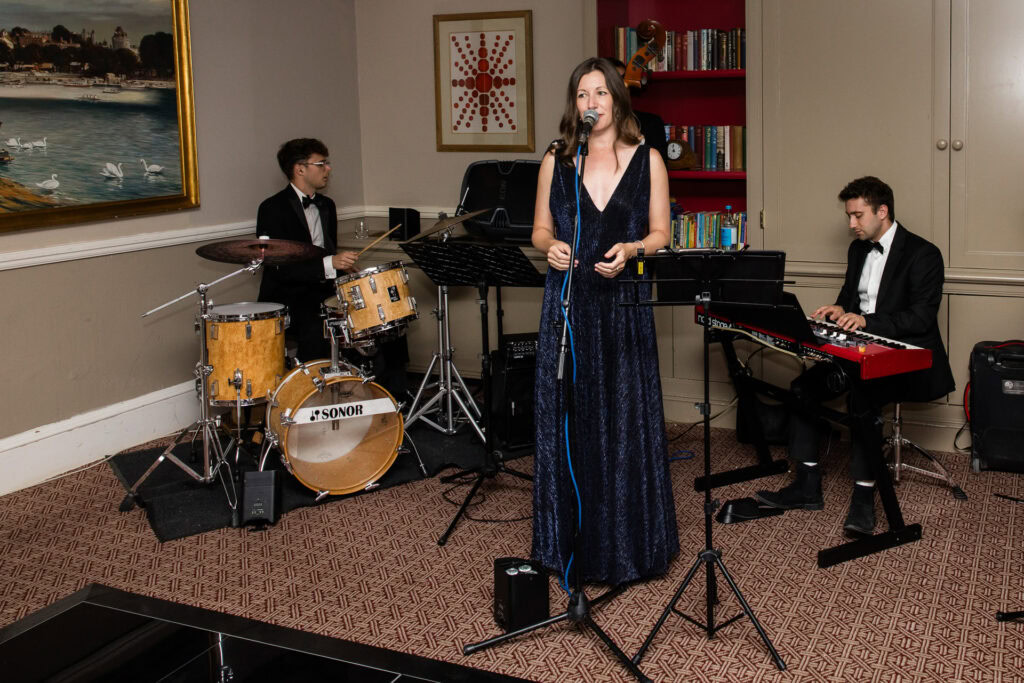 A woman sings into a microphone while two men accompany her on drums and keyboard in a cosy indoor setting with bookshelves and framed art. David Bostock Photography