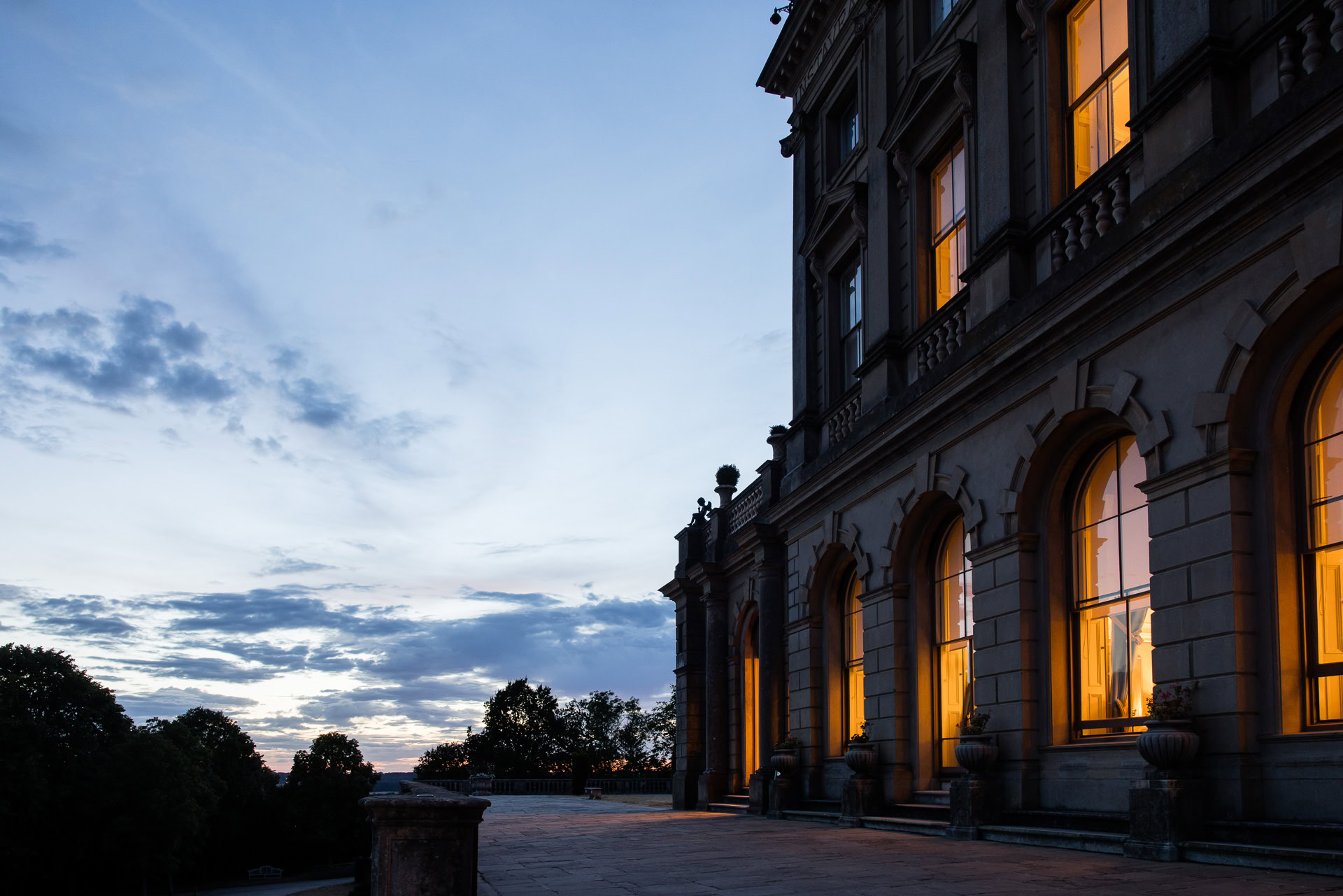 A grand building with large arched windows glows with warm light at dusk, contrasting with the deep blue sky and silhouetted trees outside. David Bostock Photography